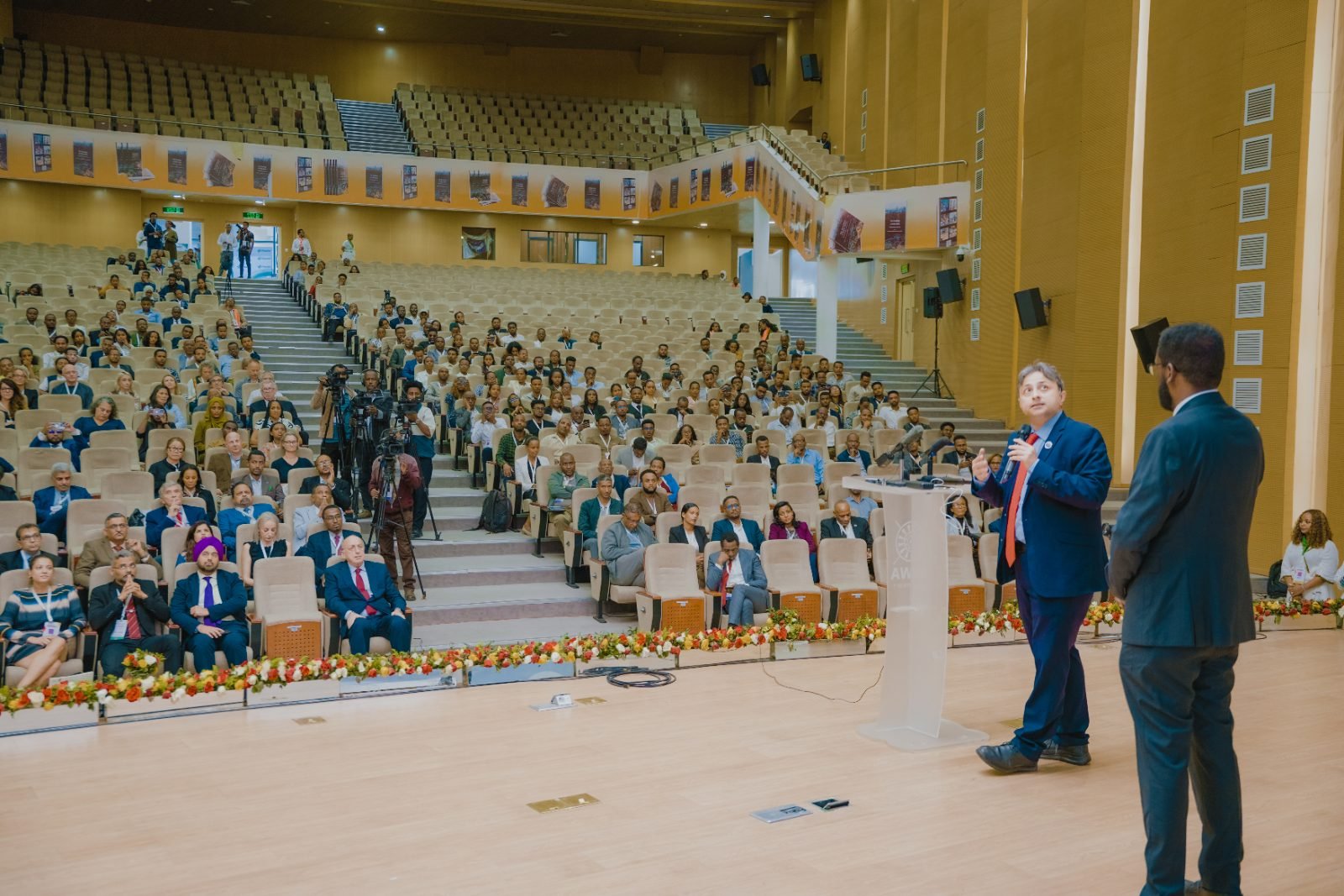 Wide view of a warm-toned auditorium filled with seated delegates. In the foreground a man in a navy suit and red tie speaks into a microphone beside a clear podium, with another man in a dark suit nearby. A garland of orange, yellow and white flowers lines the front of the stage, camera operators.