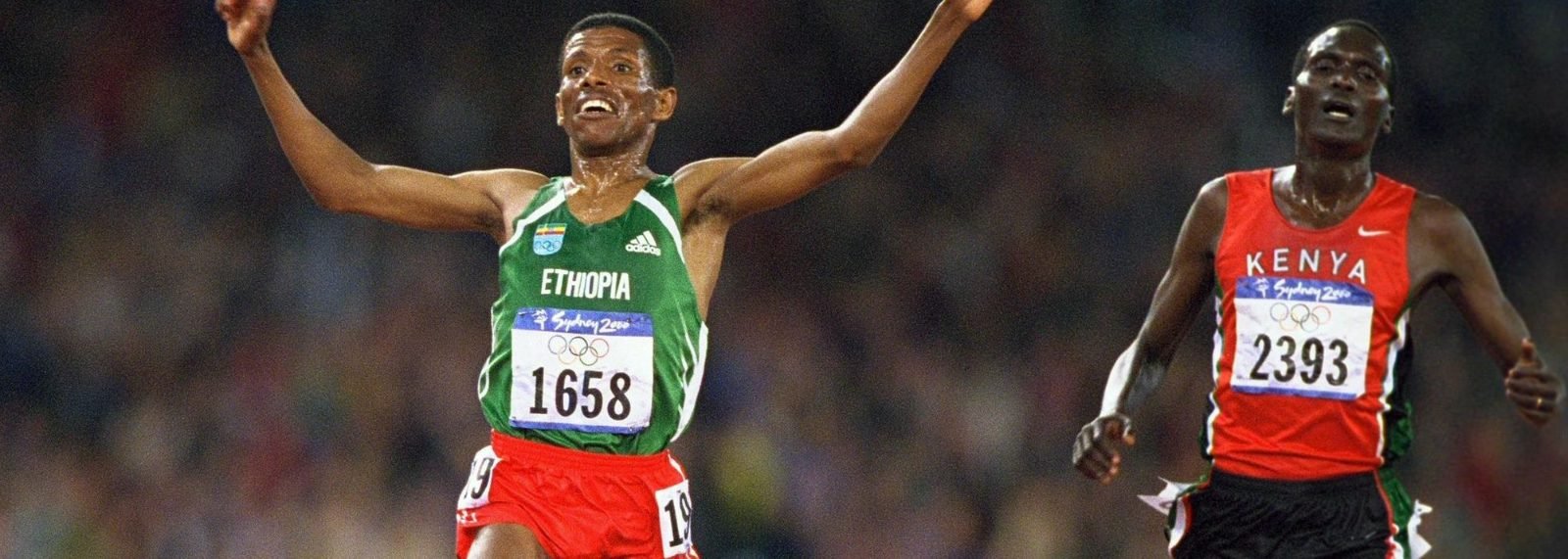 Haile Gebresillasie in green, arms raised, smiles as he crosses the line. A Kenyan runner in red trails just behind, looking tired. Both wear Sydney 2000 bibs against a blurred crowd.
