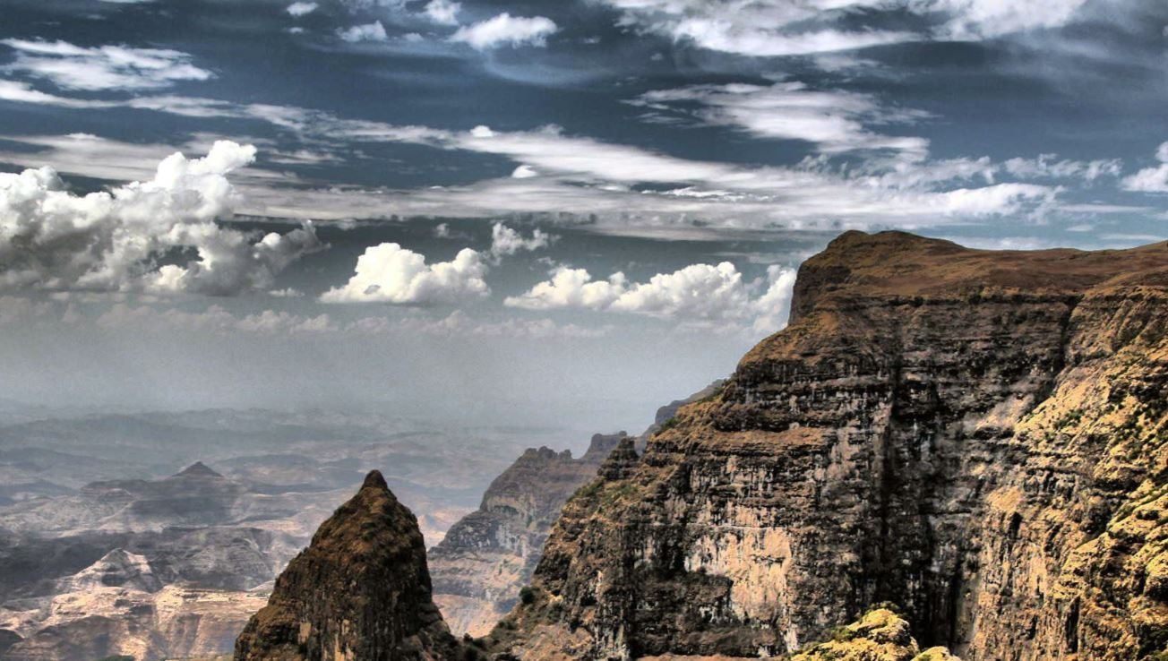 A high-contrast landscape view showing the rugged escarpments and basalt rock formations of Simien Mountains National Park in Northern Ethiopia under a dramatic, cloud-filled sky.
